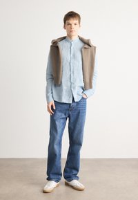 Helles blaues Leinenhemd, brauner Strickpullover über die Schultern drapiert, blaue Denim-Jeans, weiße Sneakers mit beige Sohlen.