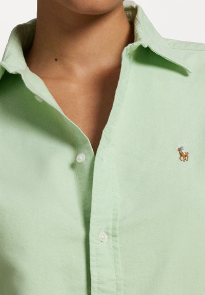 Chemise boutonnée vert clair avec une texture douce, arborant un petit logo de cheval brodé marron sur la poitrine.