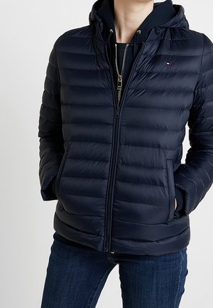 Navy blue quilted jacket with hood, front zipper, side pockets, worn over a black hoodie and paired with dark blue jeans.