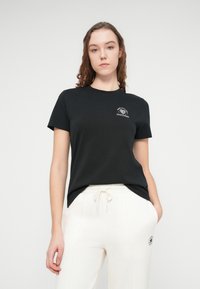 Svart bomulls-T-skjorte med korte ermer og rund hals, med hvit grafisk logo på venstre brystområde. Glatt tekstur og avslappet passform.