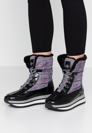 Plaidowe wysokie zimowe buty z czarnym futerkiem w środku, czarne ochronne noski, szaro-białe paskowane podeszwy, noszone z czarnymi legginsami.