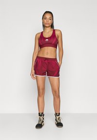 Tumši sarkans sporta krūšturis ar ģeometriskiem rakstiem, saskaņotas tumši sarkanas šorti ar baltām detaļām un melni augsti sporta apavi.