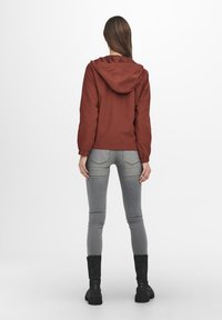 Bordově červená bunda s kapucí a elastickými manžetami, nošená přes šedé slim-fit kalhoty a černé kotníčkové boty, zobrazená zezadu. Jednoduchý design, matný povrch.