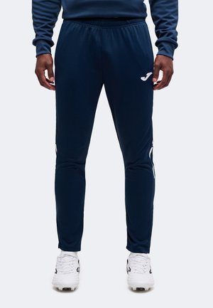 Homme portant un pantalon de sport bleu marine avec une taille élastique et des crampons blancs, debout, les mains détendues le long du corps.