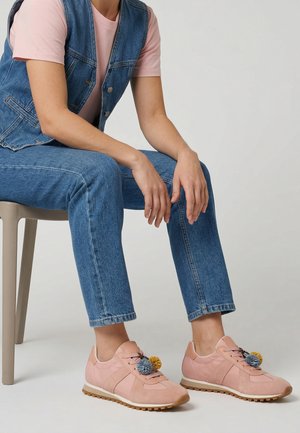 Person sitzt, trägt blaue Jeans, eine Jeansweste, ein pinkes T-Shirt und pinke Sneakers mit bunten Pompons an den Schnürsenkeln.