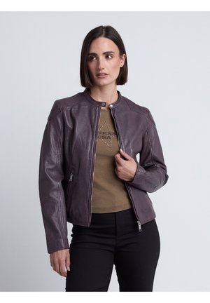 Kvinde med kort mørkt hår iført en lilla læderjakke over en brun t-shirt og sorte bukser, der kigger let til siden.
