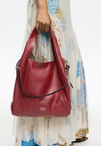 Ročna držanje velike rdeče usnjene torbe BIMBA Y LOLA nad barvno vzorčasto dolgo krilo.