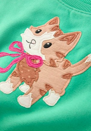 Grüner Stoff mit einer gestickten Cartoon-Katze, die ein weißes Gesicht, einen braunen Körper, eine pinke Schleife und weiße Pfoten hat, vor einem einfarbigen Hintergrund.