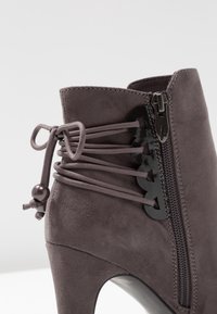 Gros plan sur une botte cheville à talon haut en suédine grise, avec une fermeture éclair sur le côté et des lacets élastiques décoratifs noués avec des perles à l'arrière.