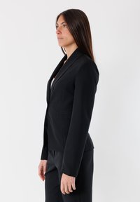 Blazer aderente nero con un design fitted, tessuto liscio, rever a punta e tasche laterali, abbinato a pantaloni neri coordinati.