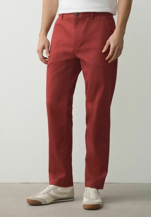 Mann trägt rostrote Straight-Leg-Hosen mit Vordertaschen, kombiniert mit weißen Sneakers und einem weißen Hemd, stehend auf einem grauen Boden.