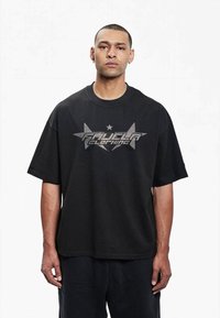 Sort oversized t-shirt i bomuld med korte ærmer. Har et sølv grafisk logo-design og en stjerneaccent på brystet.