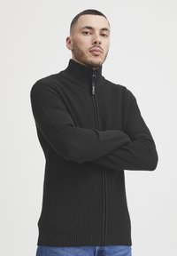 Svart hettegenser med høy krage, teksturert strikket stoff, ribbede ermer og kant, samt en frontglidelås med logo-detall.