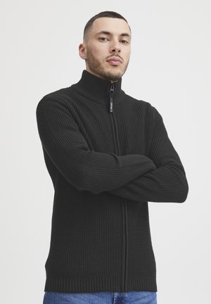 Sudadera negra con cremallera y cuello alto, fabricada en tejido de punto texturizado, con puños y dobladillo acanalados, y una cremallera frontal que presenta detalles del logo.