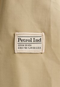Material bej cu o textură de plasă, având cusut pe el un etichetă rectangulată. Eticheta prezintă inscripția "Petrol Ind" cu text suplimentar dedesubt.