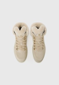 Des baskets montantes beiges avec doublure en fourrure, œillets et lacets dorés, et une surface texturée, sur un fond blanc uni.