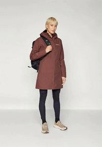 Καφέ αδιάβροχο παλτό με κουκούλα, μπροστινό φερμουάρ και πλαϊνές τσέπες. Φορεμένο πάνω από μαύρα κολάν και αθλητικά παπούτσια, συνοδευόμενο από μαύρο σακίδιο.