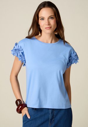 Vrouw die een lichtblauwe top draagt met bloemige kanten mouwen, grote ronde armbanden en donkere blauwe spijkerbroek, staand tegen een effen beige achtergrond.