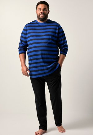 Erwachsener Mann steht barfuß, trägt ein blau-schwarz gestreiftes Langarmshirt und schwarze Hose vor einfachem Hintergrund.