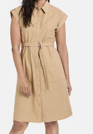 Kobieta ubrana w beżową sukienkę na guziki z kołnierzykiem, podwiniętymi rękawami typu cap, przednimi kieszeniami oraz paskiem z tego samego materiału w talii.
