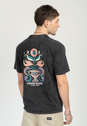 Man draagt een zwart gewassen T-shirt met een kleurrijk bloemenvaasontwerp en de tekst "Virgin Island Kaotiko Society" op de achterkant, beige broek en zilveren ketting.