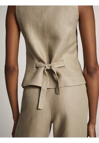 Vista posteriore di una persona che indossa un top senza maniche beige con una cintura annodata in vita e pantaloni beige coordinati.