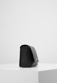 Černá kožená kabelka s klopou a stříbrným sponou, vystavená na bílém podstavci proti šedému pozadí.