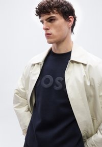 Νεαρός άνδρας με σγουρά μαλλιά που φοράει μπεζ παλτό πάνω από ένα σκούρο φούτερ με ανάγλυφη επιγραφή "BOSS", στέκεται μπροστά σε ένα απλό φωτεινό φόντο.