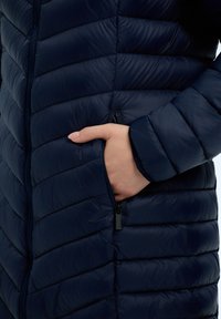 Merijärkänt tilkkuvuori, jossa on vetoketjulliset sivutaskut ja pehmeä, kiiltävä rakenne. Takki on keskikokoinen ja muotoilu on istuva.