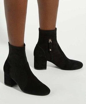 Femme portant des bottines en daim noir avec talons blocs et fermetures éclair latérales sur fond blanc.