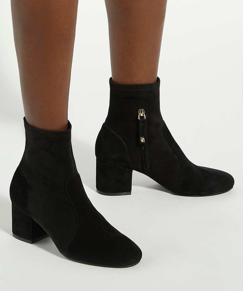 Femme portant des bottines en daim noir avec talons blocs et fermetures éclair latérales sur fond blanc.