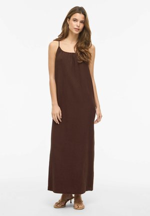 Młoda kobieta stoi, ubrana w ciemnobrązową, bezrękawnikową sukienkę maxi i beżowe sandały na obcasie na jednolitym białym tle.