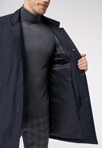 Cappotto navy con una texture liscia, rever a punta e tasca interna con zip, indossato sopra un dolcevita grigio aderente e pantaloni a quadri.