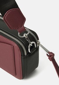 Sac rectangulaire en cuir grainé noir avec des accents bordeaux. Il dispose de fermetures éclair argentées, de quincaillerie robuste et d'une bandoulière détachable.