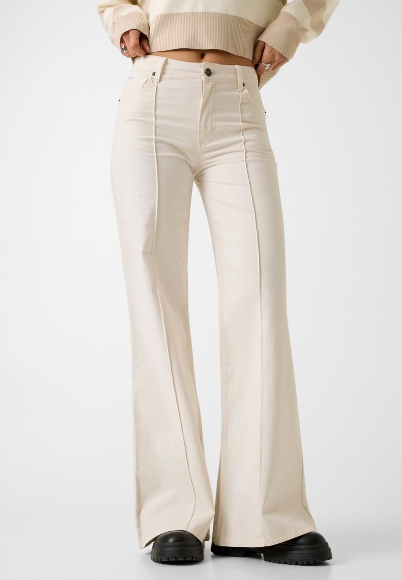 Személy, aki krémszínű, magasderekú bővülő szárú nadrágot visel elülső varrással és fekete masszív cipőkkel, a nadrág dereka felett látható egy rövid pulóver.