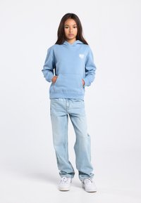 Lichtblauwe sweatvest met capuchon en voorzak, met wit "NYC" hartlogo, gecombineerd met lichte spijkerbroek en witte sneakers.