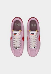 Rosa Nike-sneakers med röda detaljer, snörning och vadderad tungspets. Texturerat material med synlig branding på tungan och sidorna.