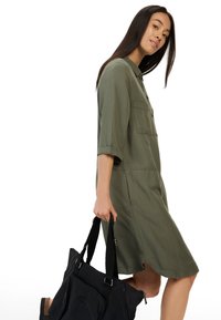 Khaki zielona sukienka koszulowa z rękawami do trzech czwarte, z guzikami z przodu i kieszeniami na piersi. Modelka trzyma czarną torbę typu tote. Gładka tekstura, swobodny styl.