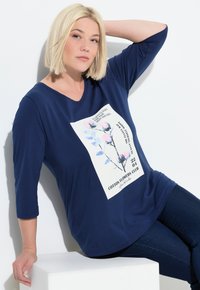 Maglietta blu navy a V con maniche a tre quarti, caratterizzata da un motivo floreale sul davanti e una vestibilità comoda. Abbinata a jeans scuri.