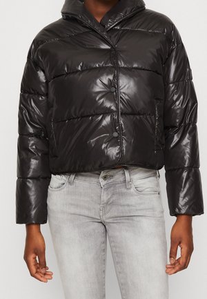 Czarna błyszcząca kurtka puchowa z przodem na zatrzaski, noszona z jasnoszarymi dżinsami, pokazująca tułów i ramiona bez widocznej głowy i stóp.