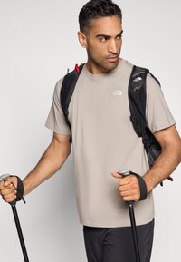 Ανοιχτό μπεζ κοντομάνικο μπλουζάκι με ένα μικρό λευκό λογότυπο, σε συνδυασμό με μαύρο σακίδιο πλάτης με κόκκινες λεπτομέρειες, κρατώντας μπαστούνια πεζοπορίας.