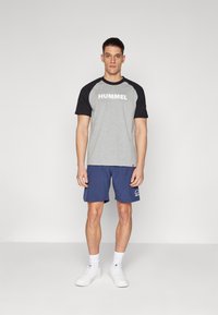 Grå t-skjorte med svarte raglanermer som har "HUMMEL" trykket i hvitt, kombinert med marineblå shorts. Hvite sneakers fullfører antrekket.