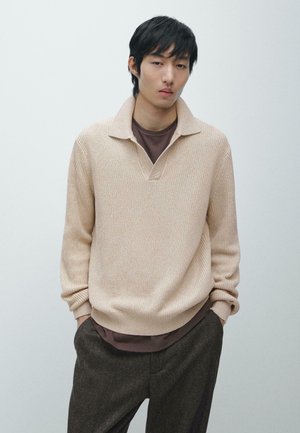 Pullover beige in maglia con collo a risvolto e apertura a V, indossato sopra una maglietta marrone, abbinato a pantaloni scuri e testurizzati.
