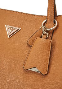 Borsa a tracolla marrone con texture, dotata di un logo triangolare in tonalità oro e un'etichetta per bagagli rimovibile con bordo metallico.