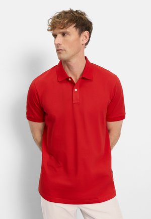 Homme aux cheveux bouclés clairs portant un polo rouge à manches courtes et un pantalon blanc, regardant vers la gauche sur un fond uni.