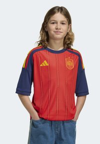 Röd och blå ungdomsfotbollströja med horisontella ränder, med Spaniens nationella emblem och gula detaljer på axlarna.