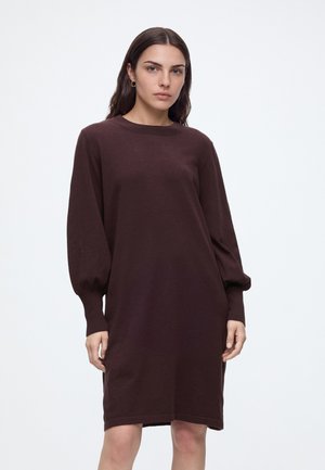 Femme aux longs cheveux foncés portant une robe marron foncé à manches longues et longueur genou, avec un col rond, debout sur un fond clair uni.
