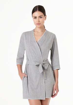 Donna che indossa un vestito avvolgente grigio chiaro al ginocchio con maniche a tre quarti, legato in vita con una cintura di stoffa, in piedi su sfondo bianco.