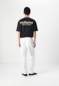 T-shirt nera in cotone con "Hollister Original Designs" in giallo sul retro, abbinata a pantaloni bianchi con vita elastica e sneakers nere.