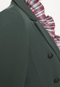 Tmavě zelený blejzr se strukturovaným designem, se skládacím, červenobílým kostkovaným límcem a dvěma černými knoflíky vpředu.